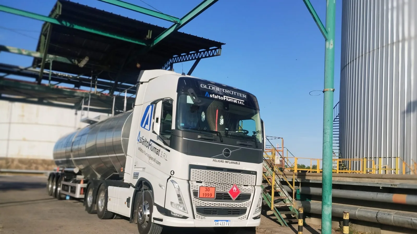 Tractor Volvo FH blanco de Asfalto Flumat con semirremolque cisterna de acero inoxidable, posicionado bajo el pórtico de carga de la planta.