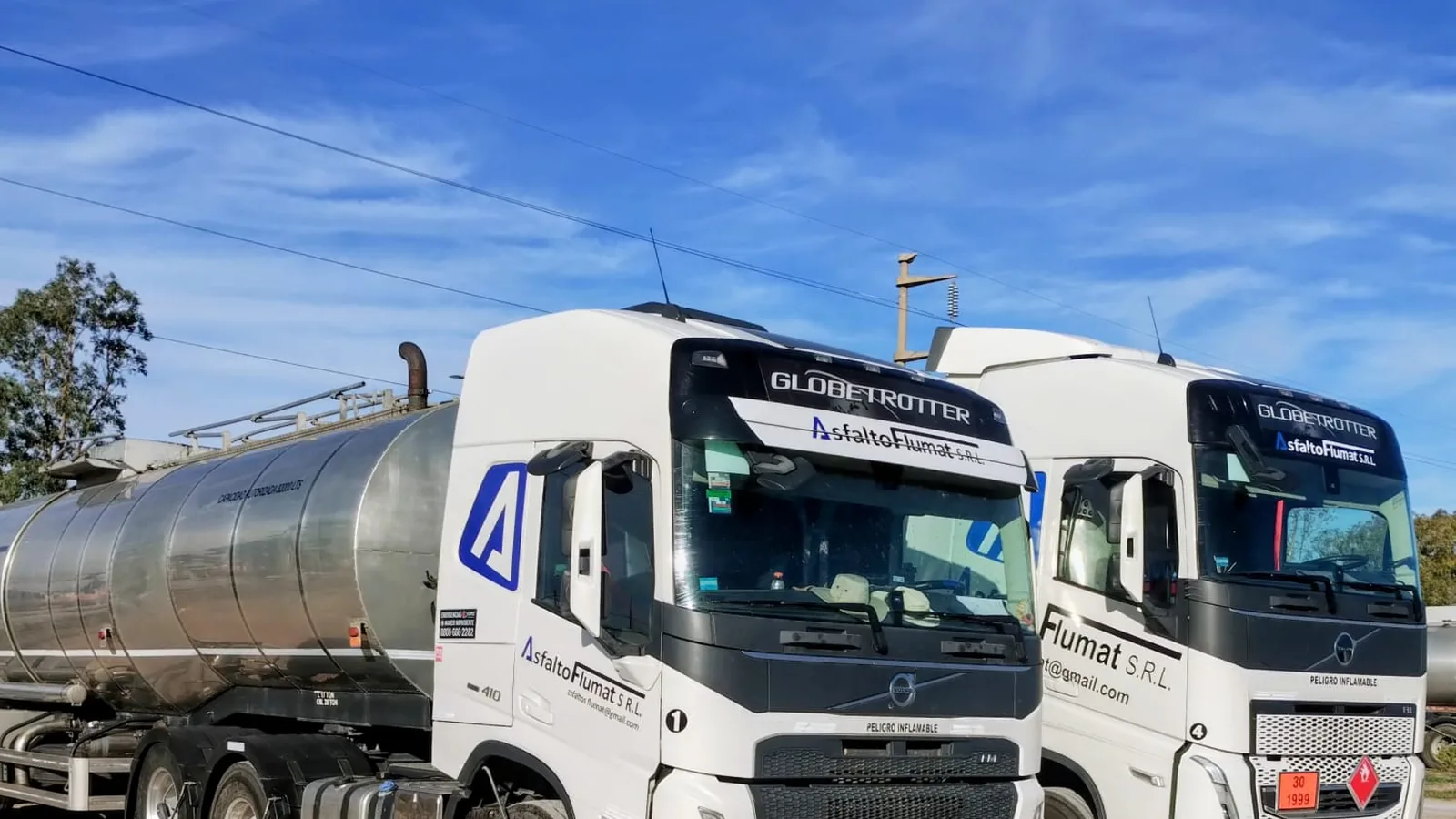 Dos tractores Volvo de Asfalto Flumat estacionados uno junto al otro con sus cisternas de acero inoxidable, bajo cielo despejado.
