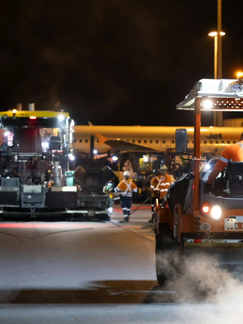 Obra nocturna de pavimentación con rodillo tándem compactando la carpeta recién colocada bajo torres de luz.