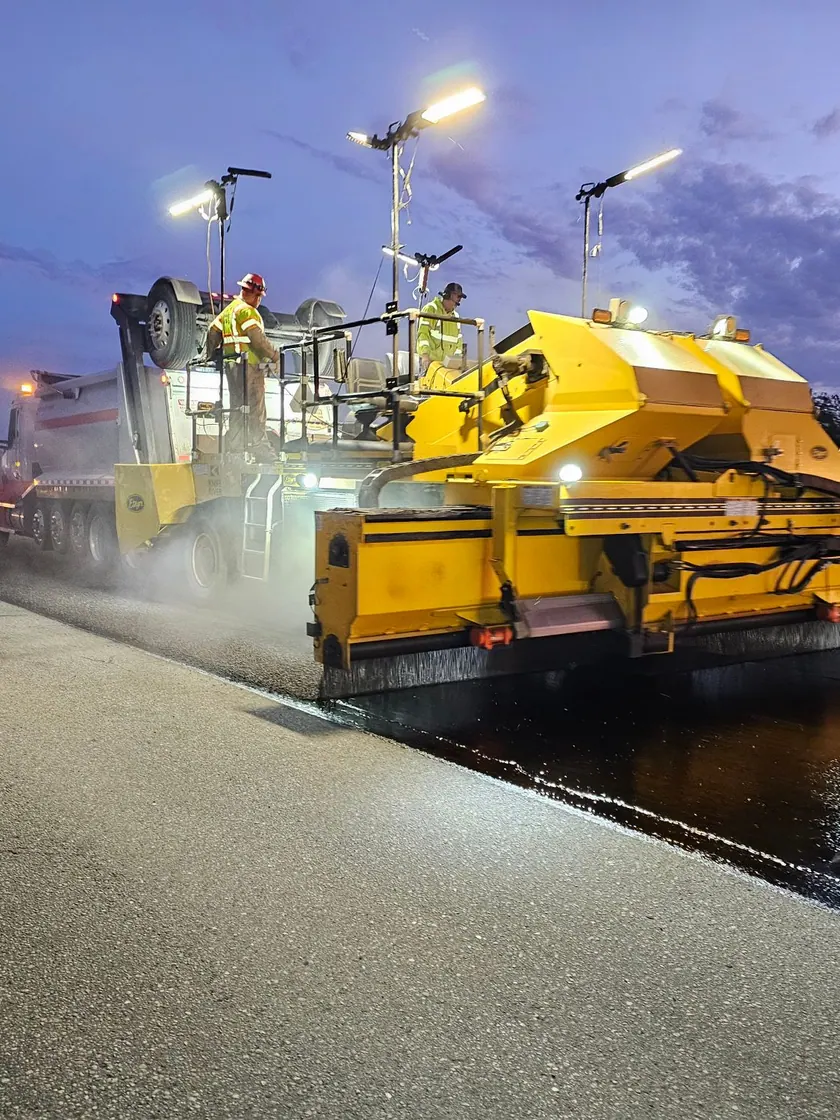 Máquina distribuidora autopropulsada aplicando ligante sobre la calzada en el crepúsculo.